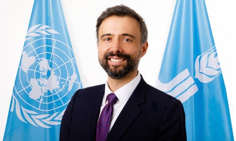 A middle-aged bearded man poses in front of blue UN flags