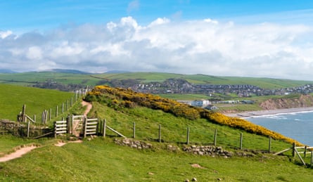 A coastal path crosses a green field over a headland with a pretty town in the distance.