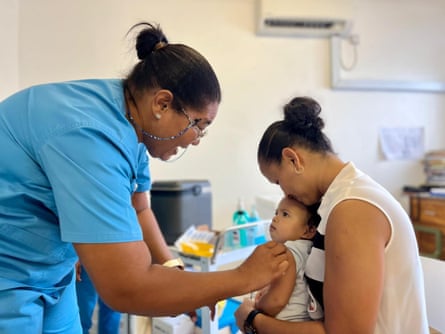 A nurse smiles at a baby being held by a woman in a medical setting. Her hand is on the baby’s upper arm