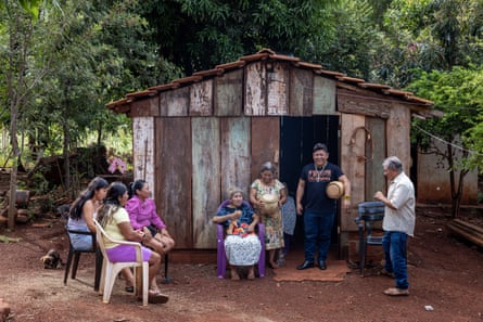 Sekelompok kecil orang berdiri, atau duduk di kursi plastik, di depan sebuah rumah kecil terbuat dari papan kayu yang lapuk.