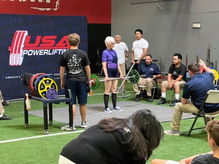 An older female lifts weights while surrounded by different athletes.