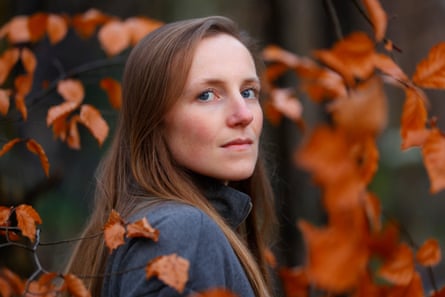A woman with long brown hair looks over her shoulder at the camera as autumn leaves fall