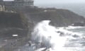 Waves as high as street lights crash over a coastal road on which cars are travelling