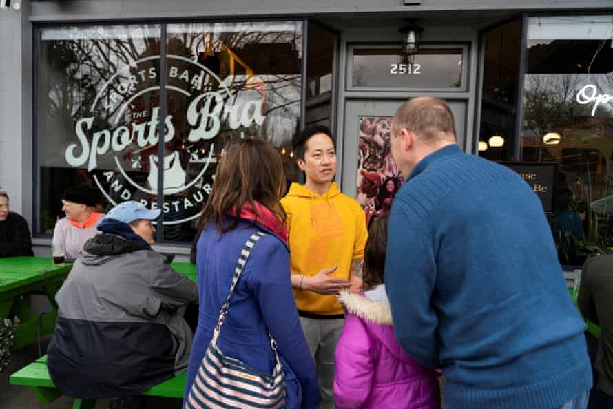A woman in a yellow hoodie talks to a family outside of a business with glass windows that have 'The Sports Bra' emblazoned on them.