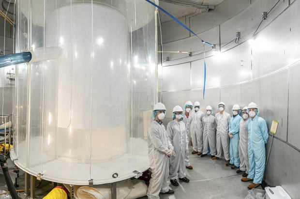A team of scientists wearing full protective suits stand in line near a device that appears to be concentric cylinders, a transparent exterior and an opaque interior, which spans from floor to ceiling.