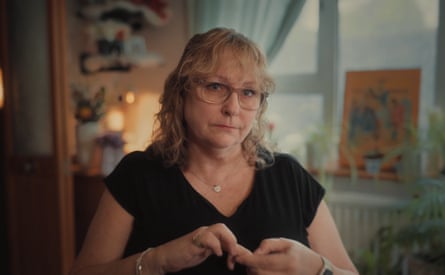 A woman with shoulder-length blond hair, light framed square glasses wearing a short-sleeve V-neck black T-shirt