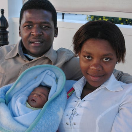 An African woman, age 18, smiles as she holds a baby in a blanket next to a young man, age 22