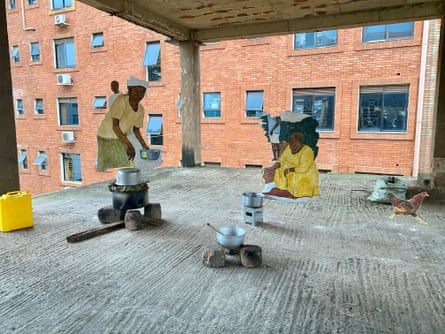 Life-sized paintings of people cooking are suspended within the concrete shell of a building over cooking pots and utensils.