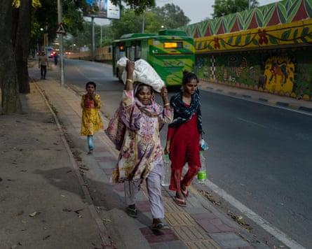 Two women and a young girl walk along a pavement, one woman carrying a bottle in each hand, the older woman carrying a bulky white sack on her head