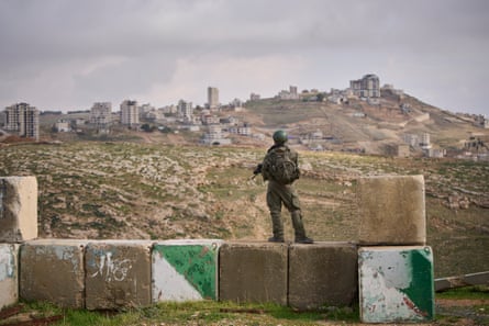 A member of the military stands on large stones holding a gun, a village in the distance