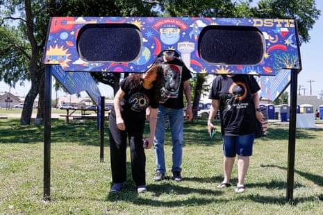 three people wearing eclipse-themed t-shirts look through a huge pair of glasses, about 10-15 feet wide and elevated on poles