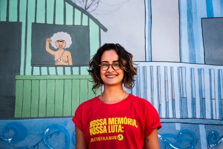 A young Latina woman in a red T-shirt stands in front of a mural of a bare-breasted woman giving a clenched-fist salute