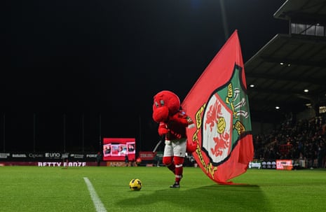 Wrexham’s mascot, Wrex the Dragon, holds a large flag whilst kicking a football.