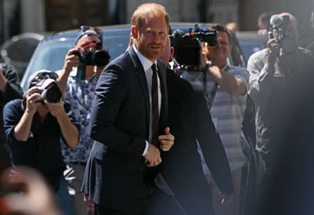 Prince Harry, wearing a suit, walks from his car surrounded by press photographers