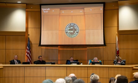 A high-ceilinged, wood-paneled room, where five people sit on an elevated space in front of a large screen.