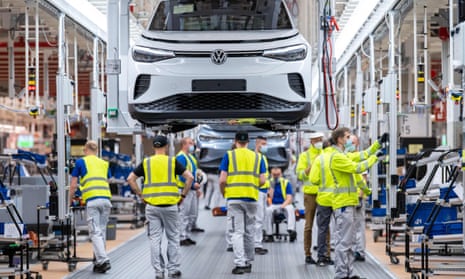 a white car hangs in the air from the production line as employees in hi-vis vests work underneath