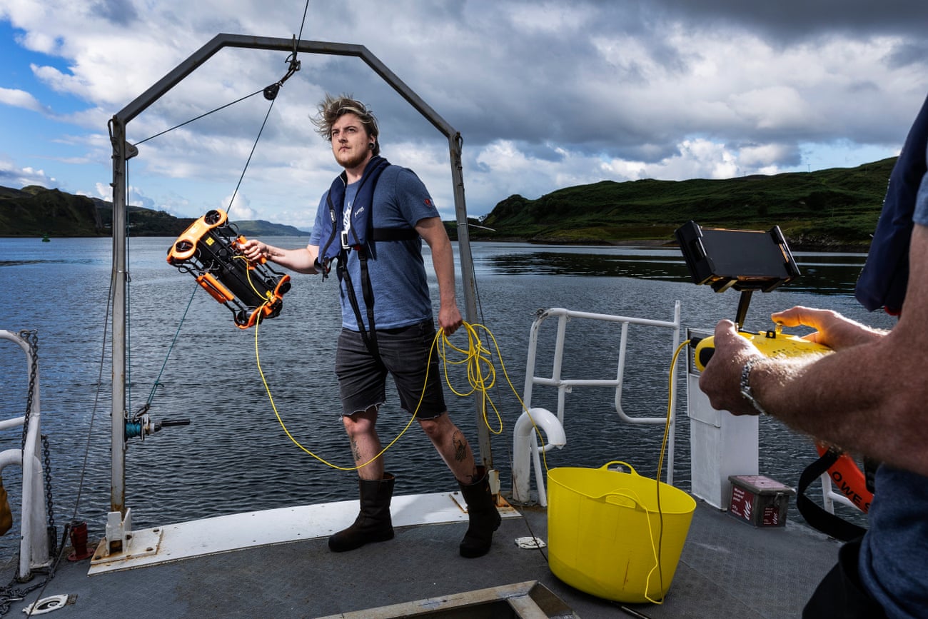 A man holds a small electrical apparatus with a cable at the back of a boat on a sea loch