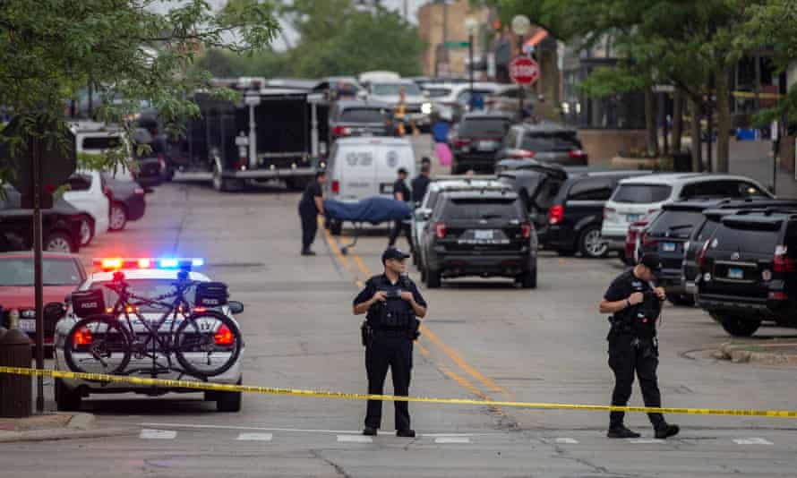 officers stand in foreground with vehicles in background and people holding stretcher