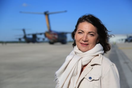 Katya Adler on an airport runway, dressed in light jacket and scarf, with planes in the background