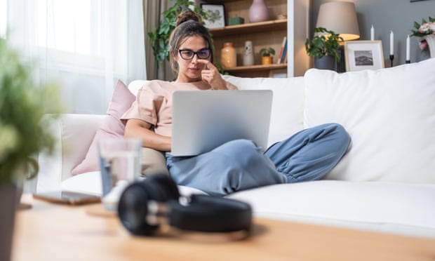 Mujer trabajando desde casa sentada en un sofá, usando un portátil. Lleva gafas y ropa cómoda, rodeada de una decoración hogareña con plantas, estanterías y cojines. En la mesa frente a ella hay unos auriculares, un vaso de agua y otros objetos personales, destacando un ambiente de teletrabajo relajado. Mujer trabajando desde casa sentada en un sofá, usando un portátil. Lleva gafas y ropa cómoda, rodeada de una decoración hogareña con plantas, estanterías y cojines. En la mesa frente a ella hay unos auriculares, un vaso de agua y otros objetos personales, destacando un ambiente de teletrabajo relajado.