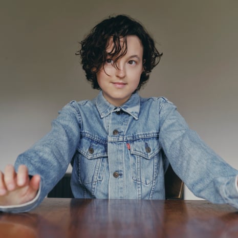 Bella Ramsey is seated, wearing a denim jacket, with the heels of her hands leaning on a round wooden table