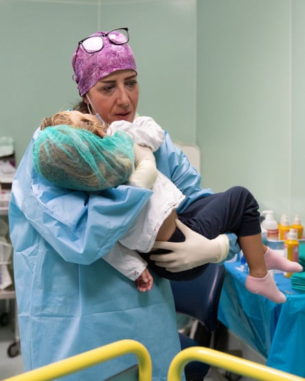 A woman in pale blue scrubs with a pink scarf over her hair holds a small girl in her arms on a hospital ward.