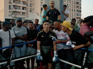Mehdi stands in the corner of a ring wearing boxing gloves surrounded by supporters