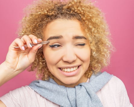 A woman is pulling out the hair from her brows using a tweezer for that. It is painful. She doesn’t like it. Isolated on a pink background. Posed by a model.