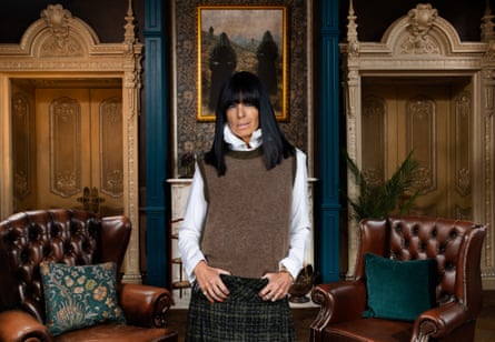 A woman with dark fringe wearing a white top, brown woolen vest and a black plaid skirt stood next to two leather chairs in front of a painting of two dark fugures