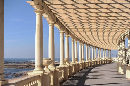 A shaded curved walkway with cream pillars next to the sea