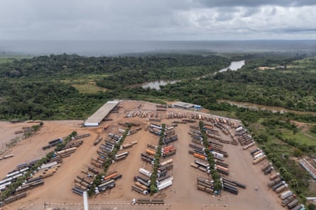 Dozens of parked trucks can be seen with the river and dense forest behind them.