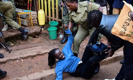 A female officer in khaki uniform stands over and grabs the arm of a young woman who is lying on her back on some steps; a person holding a sign is looking on and the legs of another officer are seen heading up the steps