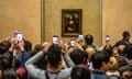 Lots of tourists using cameras and mobile phones take pictures  of the Mona Lisa behind glass in the Louvre state room.