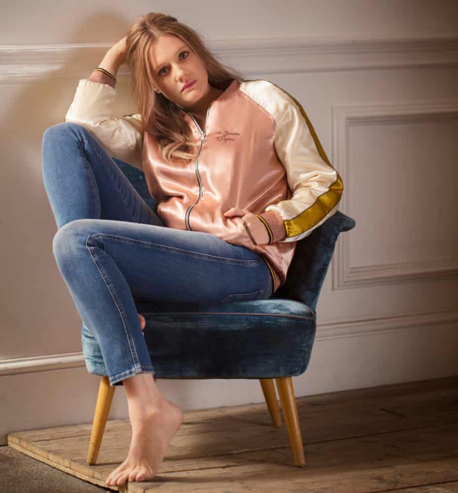 Romola Garai sitting on a chair wearing a pink bomber and jeans