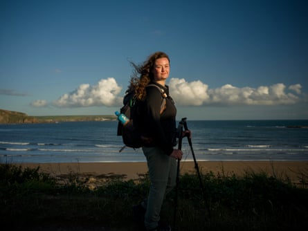 A woman holding walking poles and carrying a backpack stands with the sea behind her.