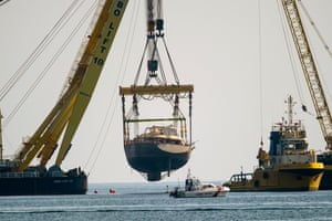 The superyacht is suspended a metre or two above the sea, hoisted by a crane and surrounded by salvage equipment