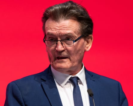 Feargal Sharkey wearing a blue suit standing at a podium in glasses. Behind him is a red wall.