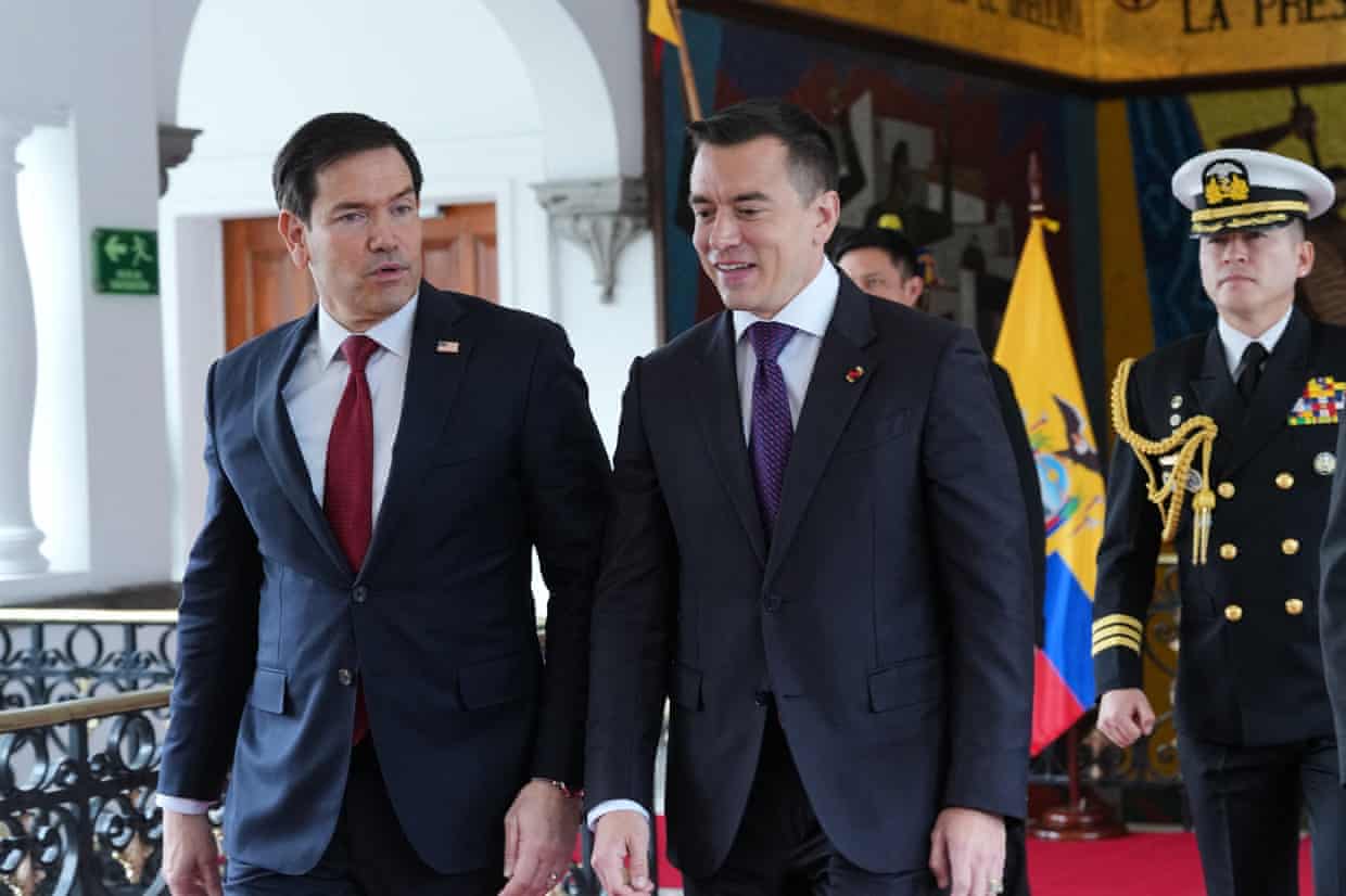 Two middle aged men in suits speak as they walk, with a man in military uniform behind them.