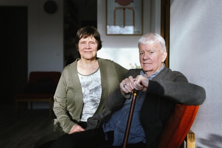 An older woman with short brown hair, wearing a green cardigan over a patterned T-shirt, sitting in a living room next to an older man with white hair, wearing a blue shirt and a grey cardigan, and holding a cane