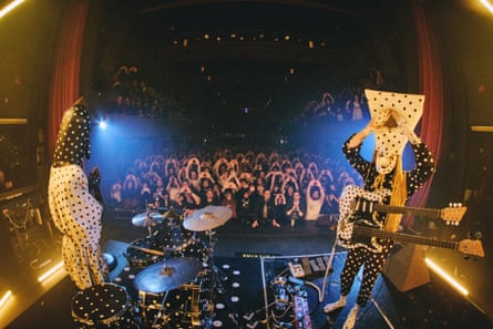 Two musicians with spotty outfits and spotty boxes on their heads in front of fans who have their arms up in the air