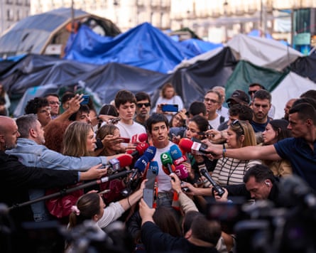 Alicia del Río addresses a crowd of journalists around her with tents in the background
