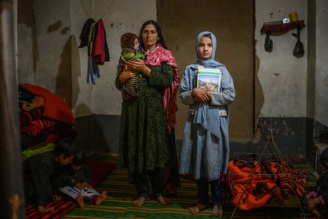 A mother holding a baby stands next to a girl in a room of a family home.