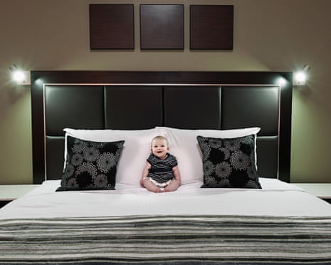 A smiling baby in a striped shirt sits propped against white pillows on a bed with black patterned cushions