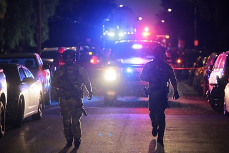 Two uniformed FBI officers are backlit with the flashing red and blue lights of emergench vehicles as they walk down a dark street lined with parked cars at night.