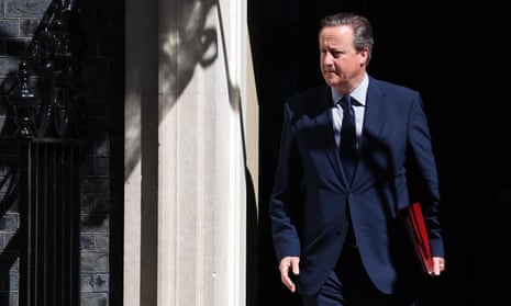 David Cameron walks out of a building holding a red folder.