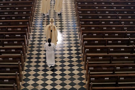 An archbishop in gowns walks through an empty church followed by an assistant, both in vestments.
