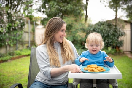 A woman serving a plate of nuggets to an enthusiastic blond-haired toddler in a high chair in a back yard