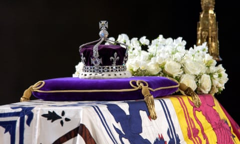 A purple and white velvet crown with metal and jewels on a purple pillow next to a mass of white flowers on top of a flag on top of a coffin.