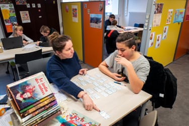 Chloé sitting at a desk beside a student, pointing at a card with text on it