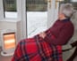 Elderly old woman at home with a rug on knees keeping warm indoors by electric fire with snow outside the windows